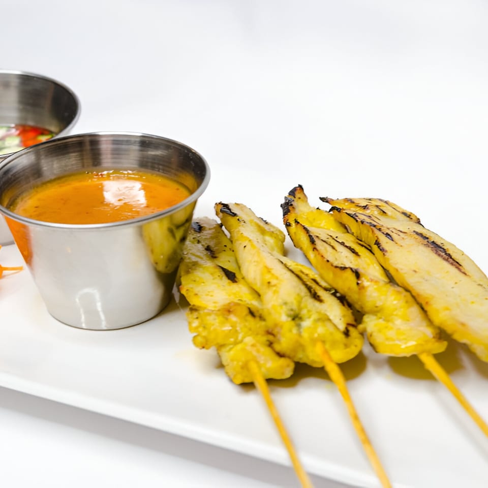 Grilled chicken satay skewers with two dipping sauces in metal cups on a white plate.