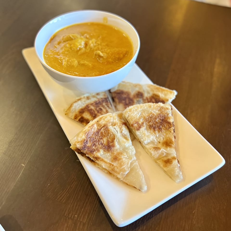 A white bowl of yellow Thai curry with chicken, served with crispy roti bread on a white plate.