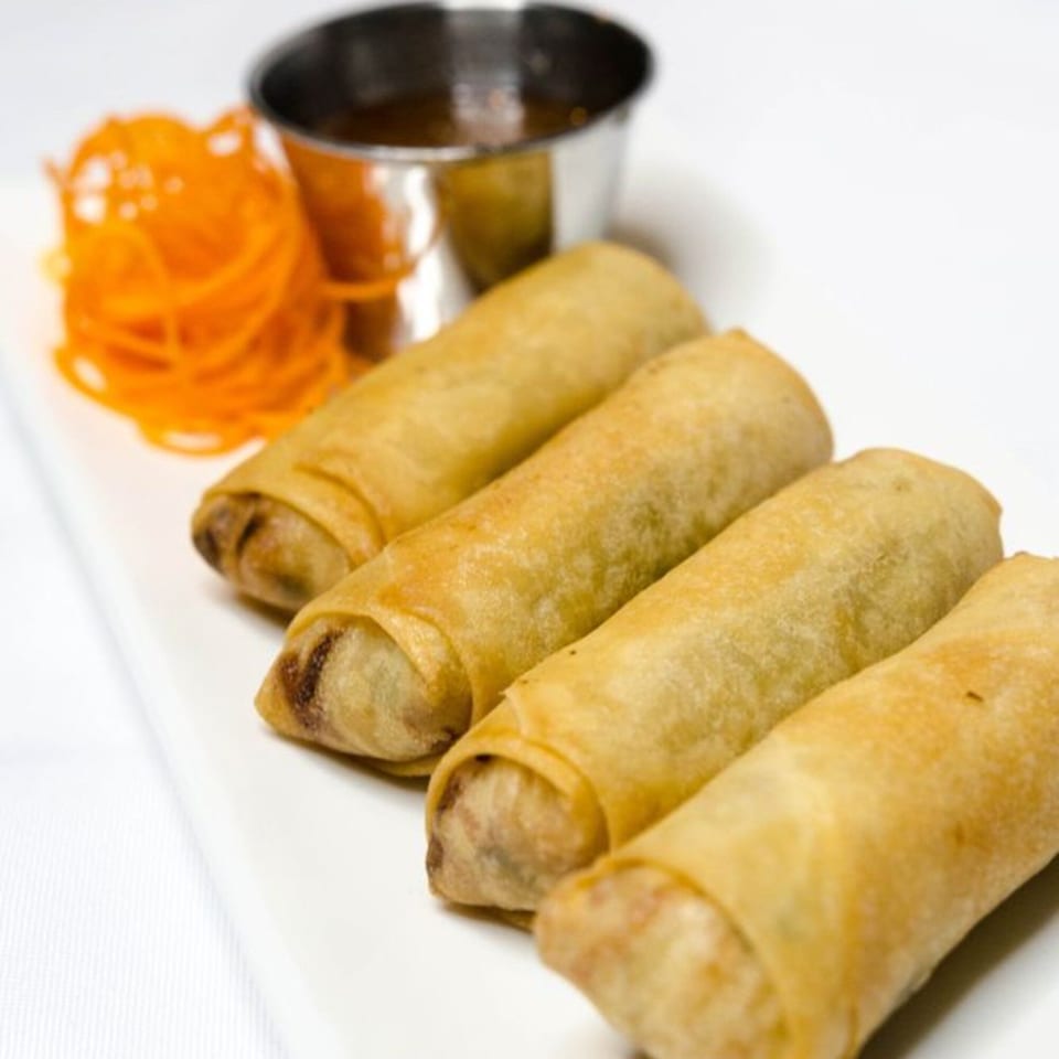 Four crispy Thai spring rolls on a white plate with shredded carrots and dipping sauce.